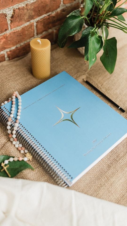 The planner on a table beside a plant, a candle and a rosary