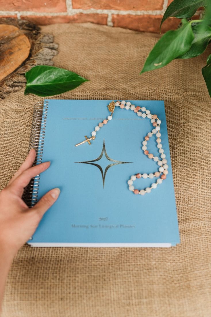 A hand resting on the blue cover of the Traditional Catholic Planner 2027, with a rosary across it
